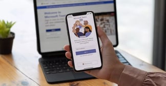 Hand holding cell phone - Microsoft Teams on screen - laptop in the background