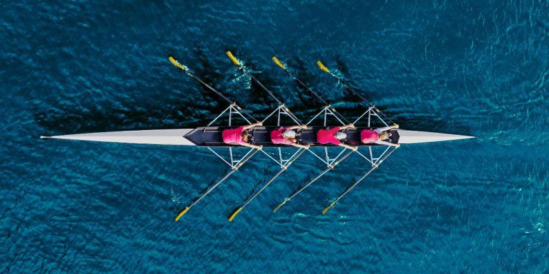 A team of four people wearing red shirts rowing a boat on water in sync, seen from above