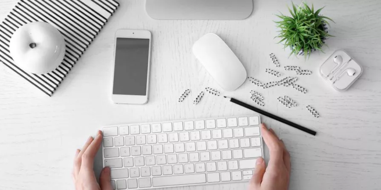 A white desktop featuring an array of Apple products including an iPhone, a keyboard, a mouse, headphones and monochrome paperclips with a green plant