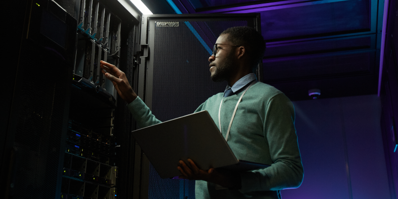 Man standing next to a server stack holding a laptop in his left hand, his right hand hovering on hard drives