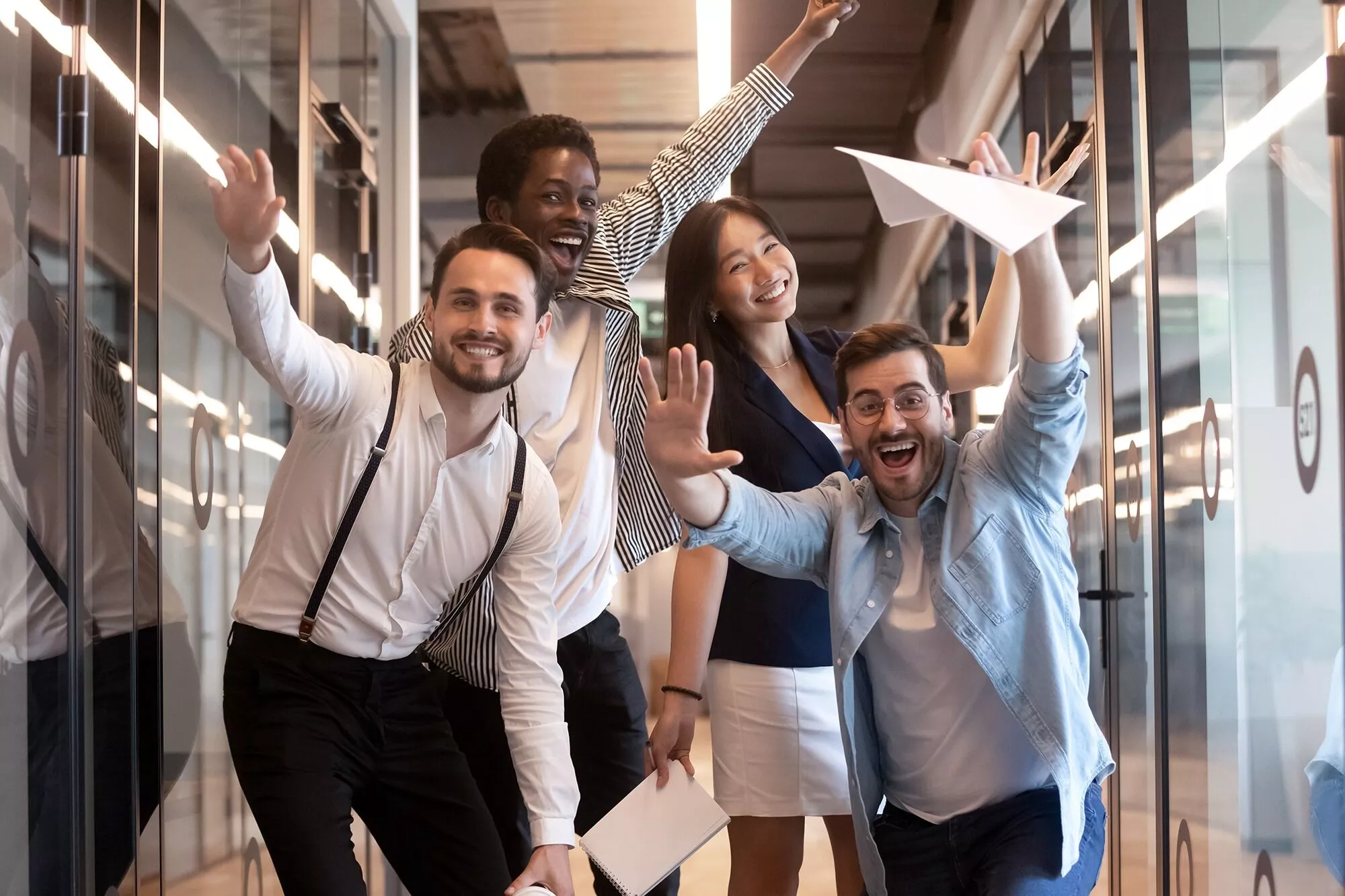 A cheerful group of four diverse office colleagues celebrating a success with raised arms and paper airplanes, in a modern office setting.